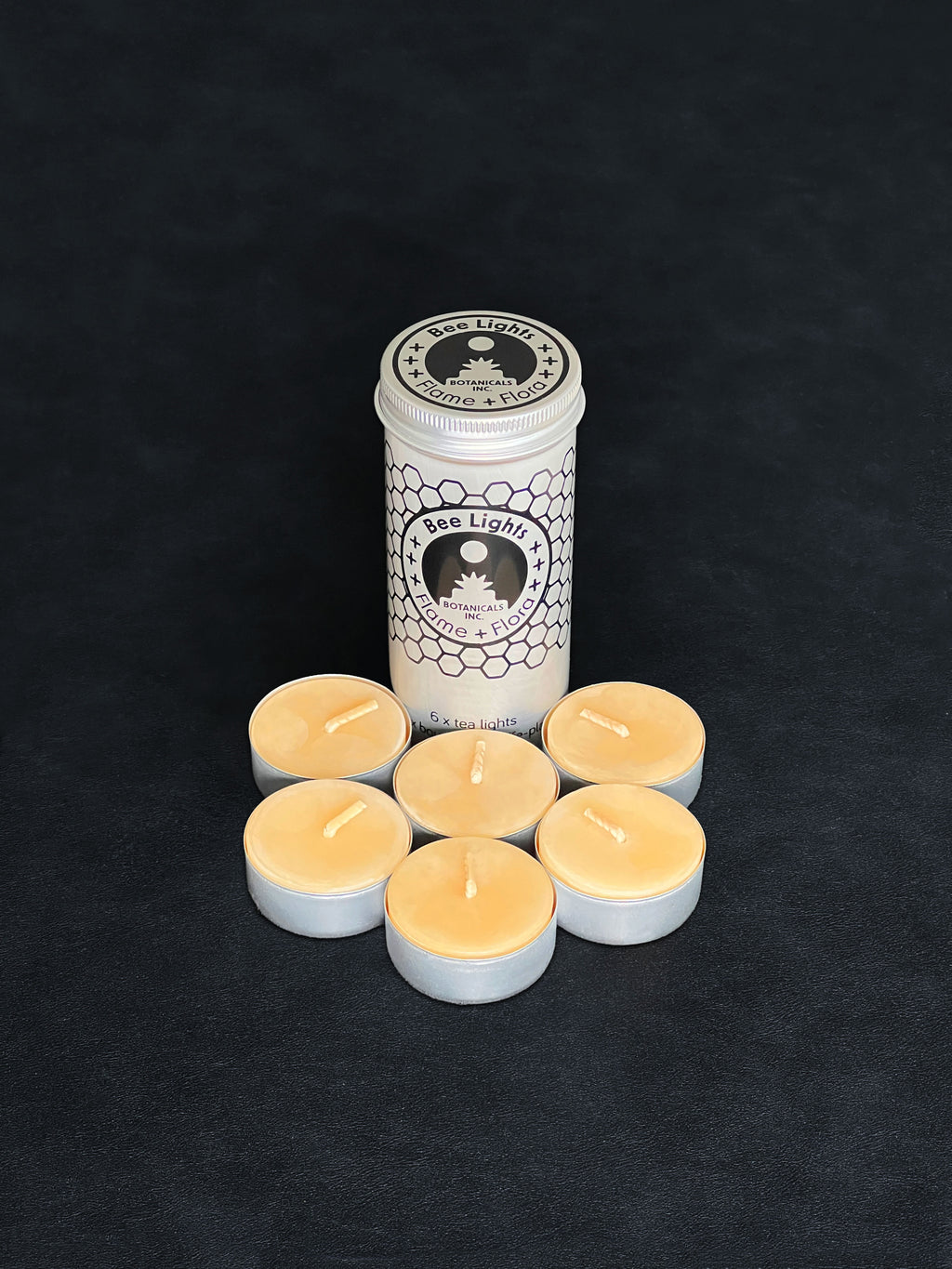 Beeswax tealight storage tin with black patterned design and six tealights with cotton wicks in metal cups displayed in the foreground on a dark background