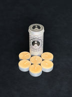 Beeswax tealight storage tin with black patterned design and six tealights with cotton wicks in metal cups displayed in the foreground on a dark background