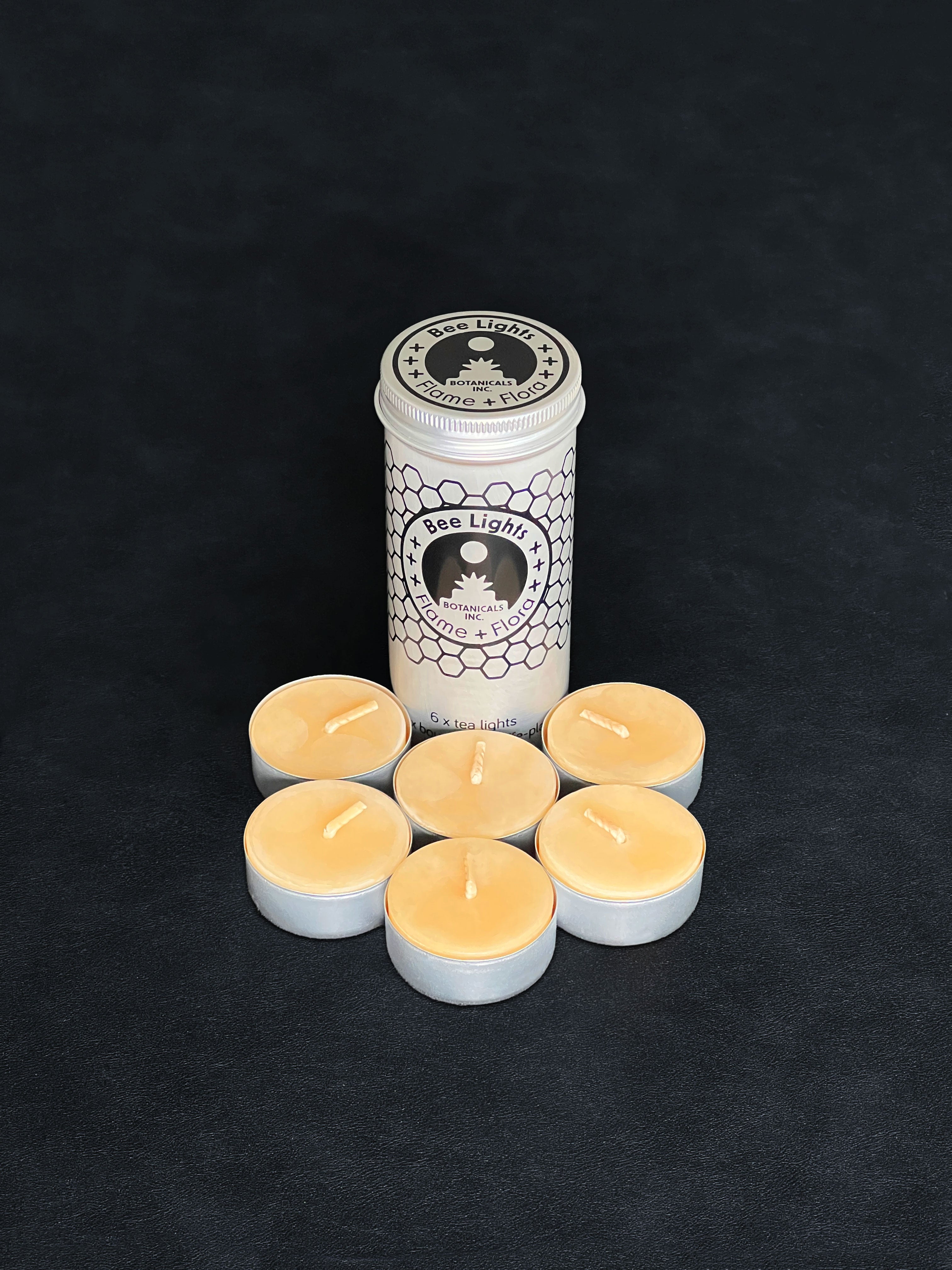 Beeswax tealight storage tin with black patterned design and six tealights with cotton wicks in metal cups displayed in the foreground on a dark background