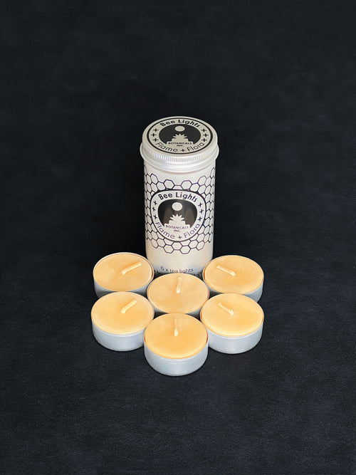 Beeswax tealight storage tin with black patterned design and six tealights with cotton wicks in metal cups displayed in the foreground on a dark background