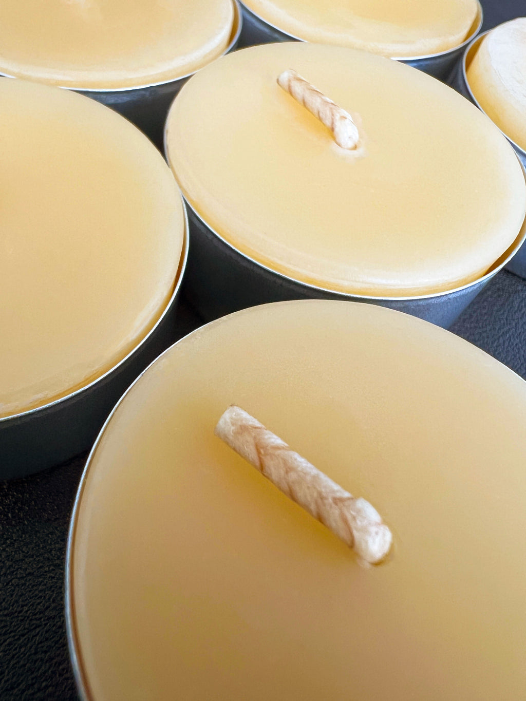 Close-up of several beeswax tealight candles in metal cups with visible cotton wicks on a dark surface