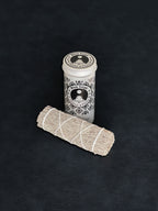 Blue Sage storage tin with black patterned design and a bundle of Blue Sage tied with white cotton string displayed in the foreground on a dark background