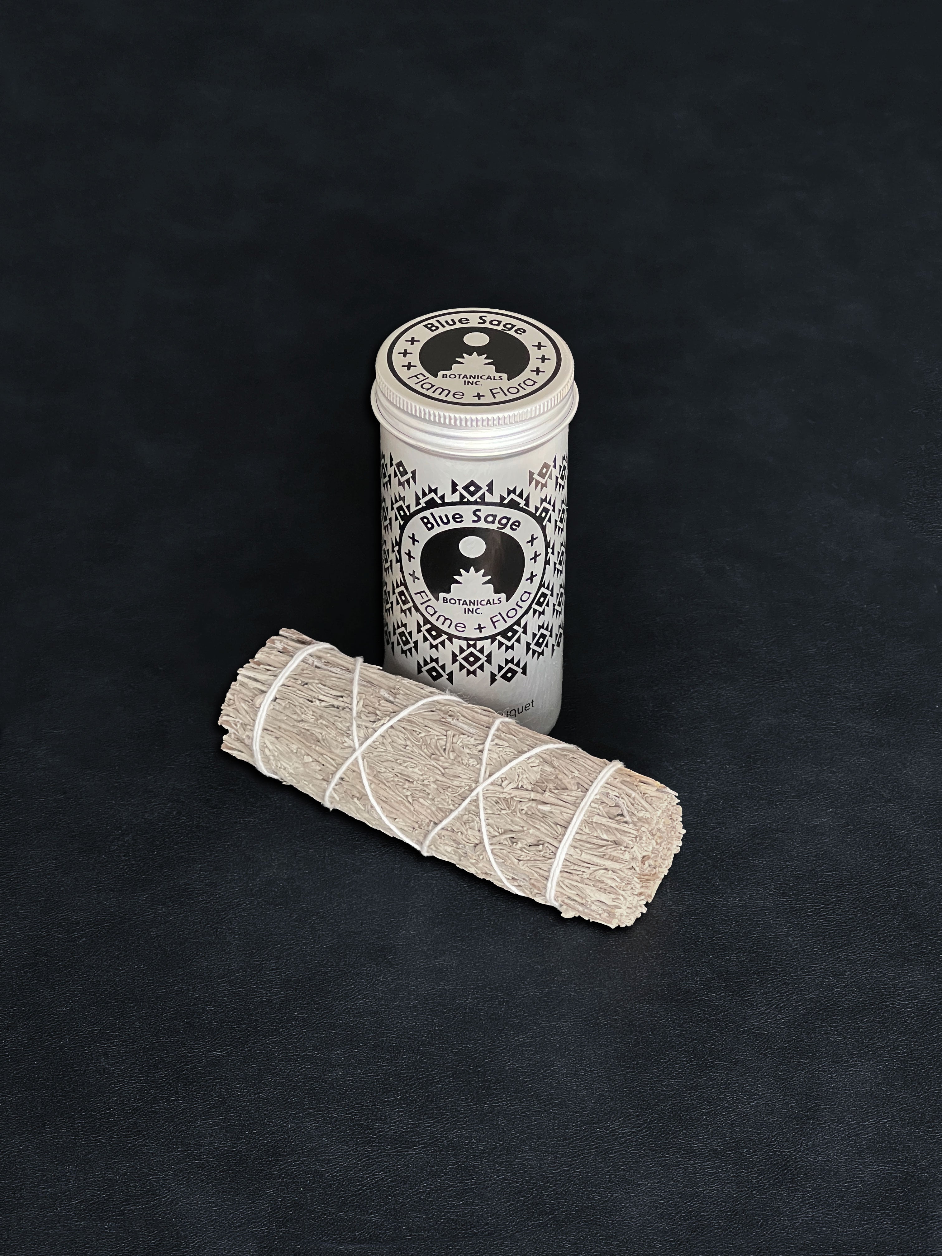 Blue Sage storage tin with black patterned design and a bundle of Blue Sage tied with white cotton string displayed in the foreground on a dark background