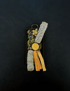 Bird’s-eye view of a rectangular black cast iron incense burner with handles on either end holding a beeswax tealight surrounded by palo santo, blue sage, white sage, juniper and eucalyptus botanicals on a dark background