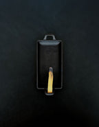 Bird’s-eye view of a rectangular black cast iron incense burner with handles on either end holding a palo santo stick with a glowing ember and rising smoke against a dark background