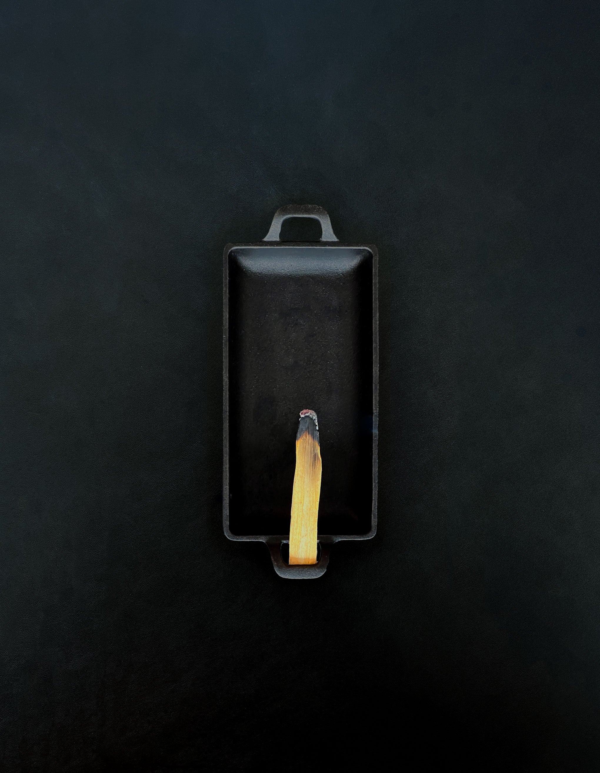 Bird’s-eye view of a rectangular black cast iron incense burner with handles on either end holding a palo santo stick with a glowing ember and rising smoke against a dark background