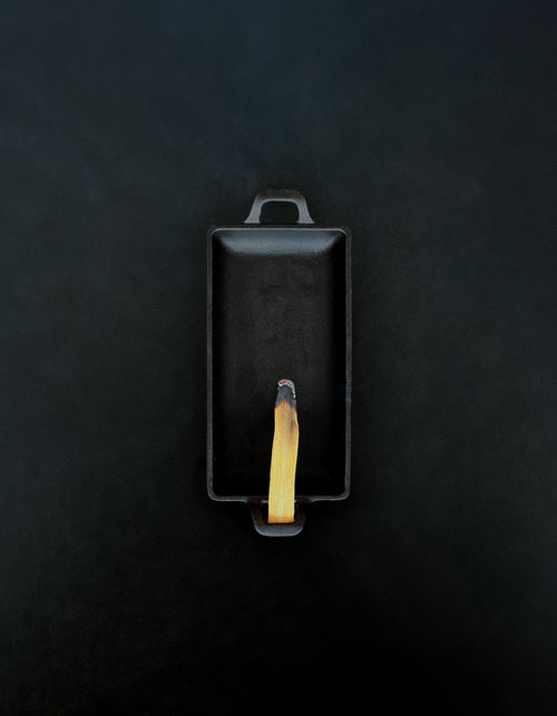 Bird’s-eye view of a rectangular black cast iron incense burner with handles on either end holding a palo santo stick with a glowing ember and rising smoke against a dark background