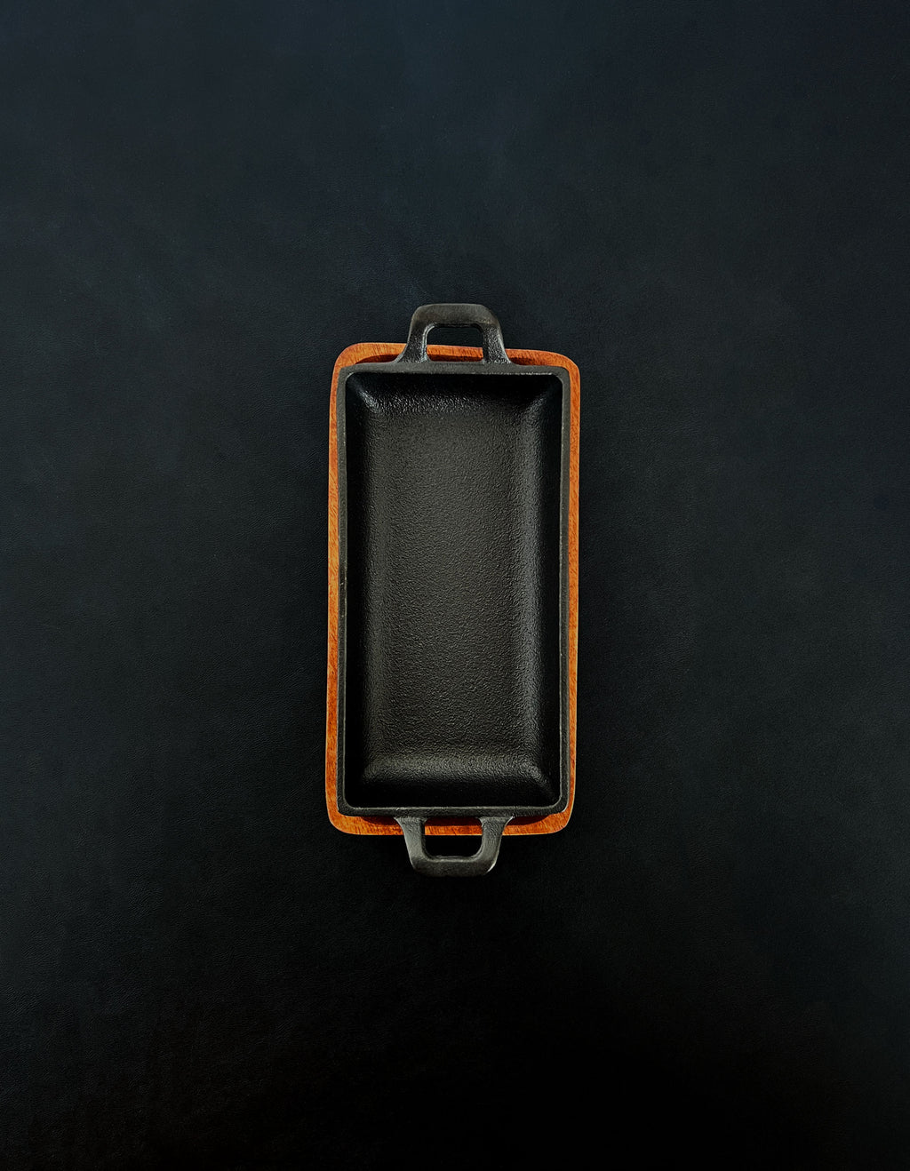 Bird’s-eye view of a rectangular black cast iron incense burner in a vertical orientation with handles on both ends, nested in an acacia wood tray against a dark background