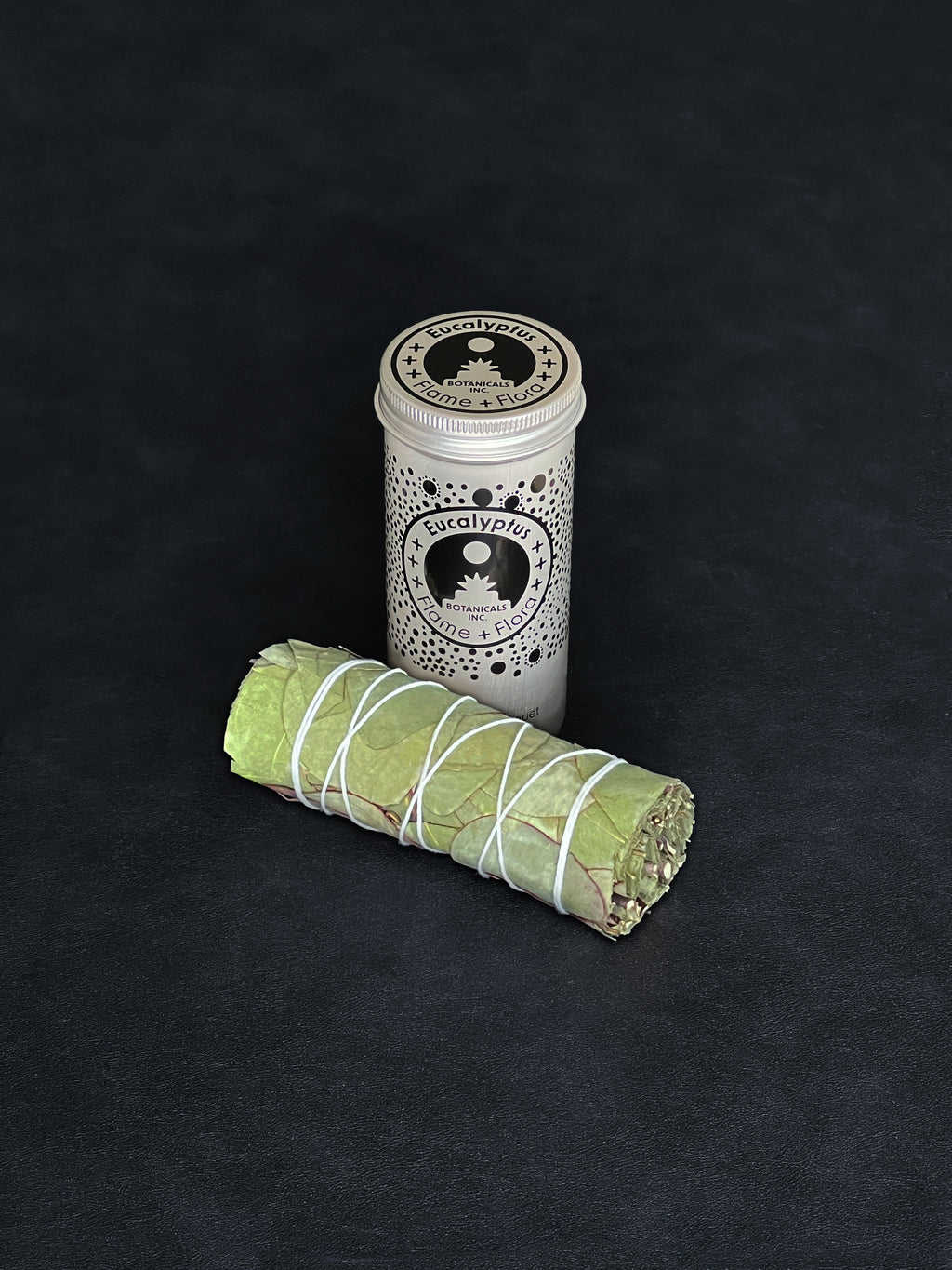 Eucalyptus storage tin with black patterned design and a bundle of Eucalyptus tied with white cotton string displayed in the foreground on a dark background