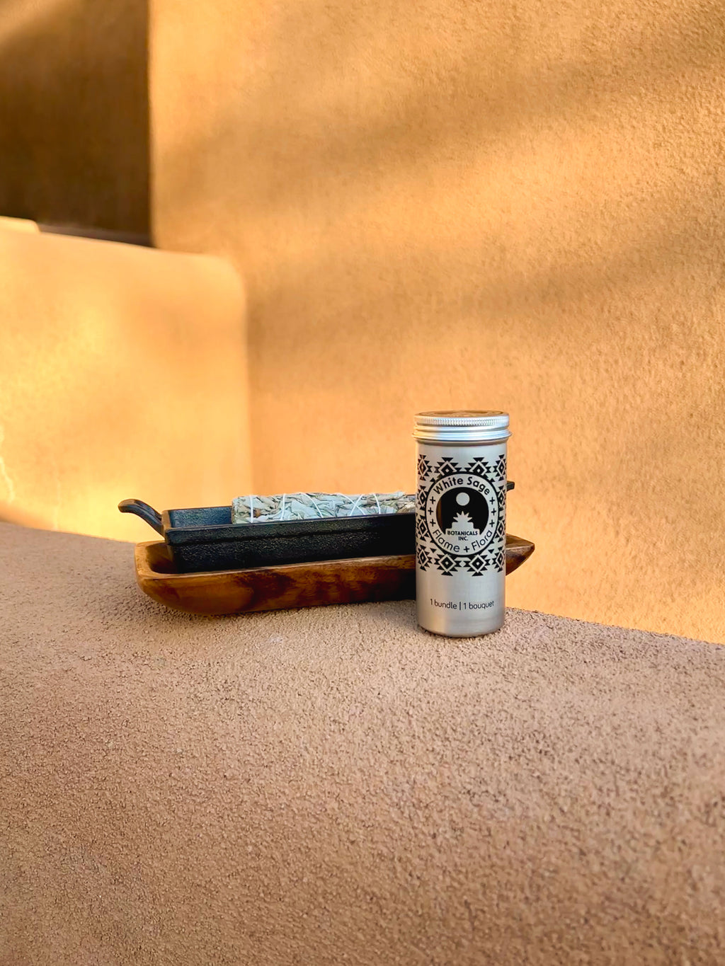 White Sage storage tin with cast iron incense burner and acacia wood tray holding White Sage tied with white cotton string on a beige adobe-style garden wall in a styled outdoor setting