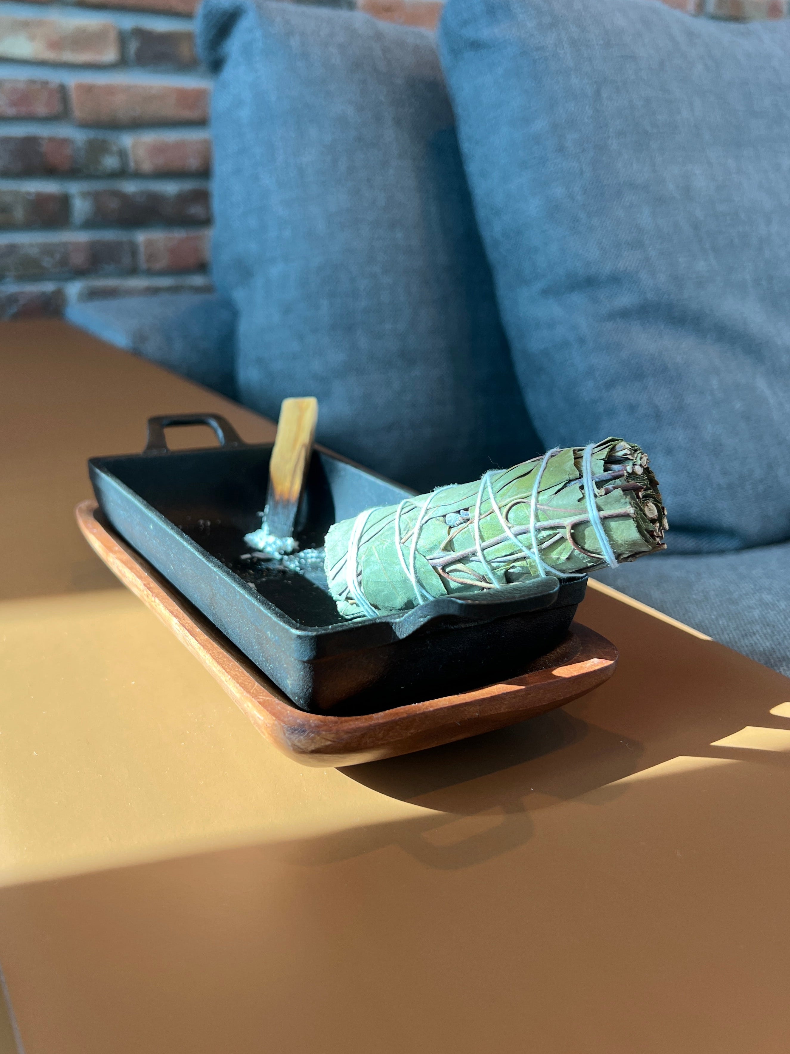 Rectangular cast iron incense burner and acacia wood tray set holding a bundle of eucalyptus and a stick of Palo Santo, sitting on a coffee table with a couch and brick wall in the background.