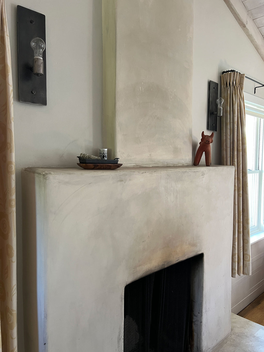Cast iron incense burner and acacia wood tray set holding a blue sage bundle, with the metal Travel Tin behind it. Resting on the mantle of a white fireplace with decorative items on a white wall with a window and curtain.
