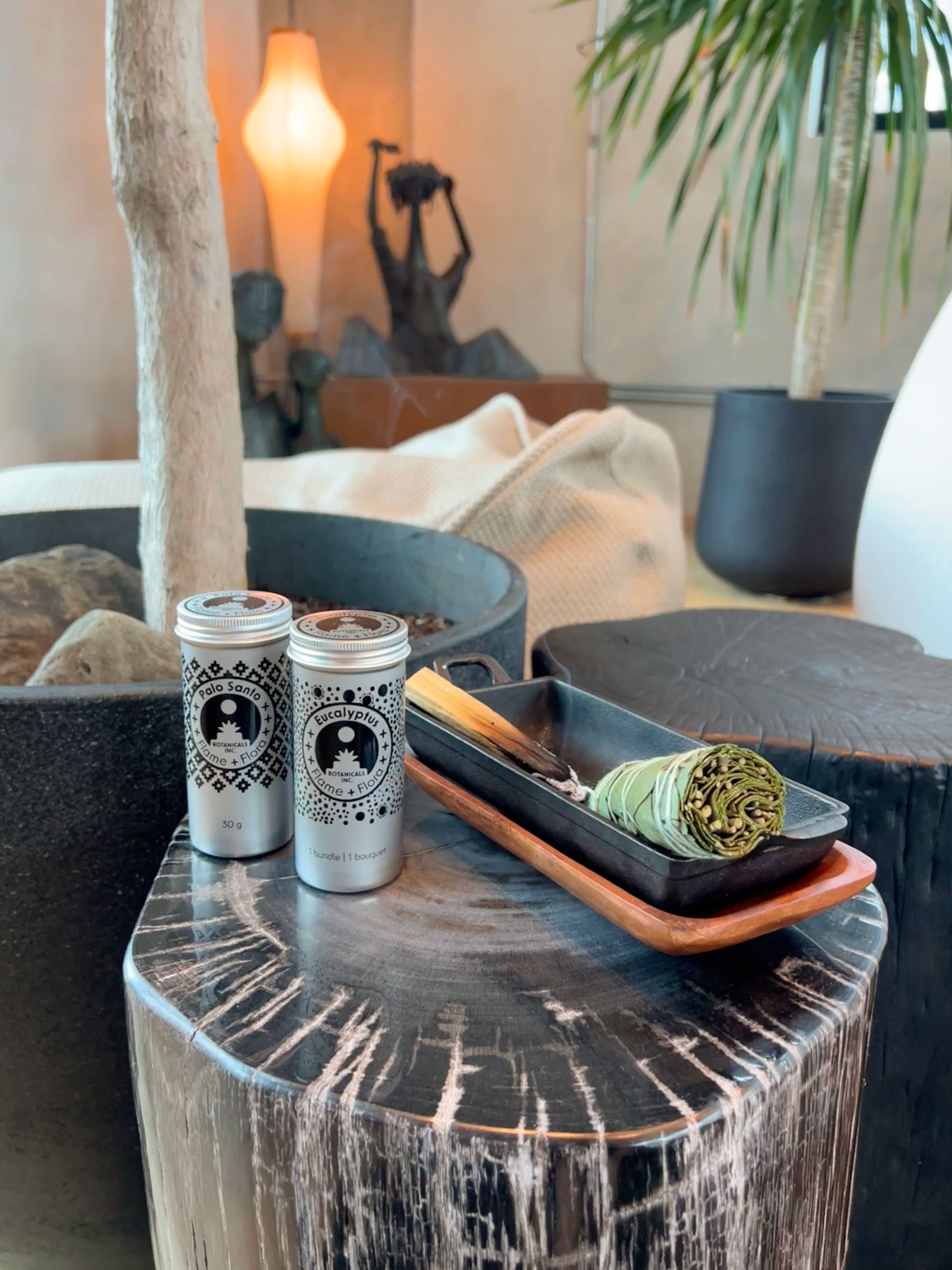 Two Flame and Flora cans with sit on a rustic dark wood side table next to a tray holding a bundle of sage smudge and a smouldering stick of Palo Santo, in a styled indoor setting with plants and warm ambient lighting.