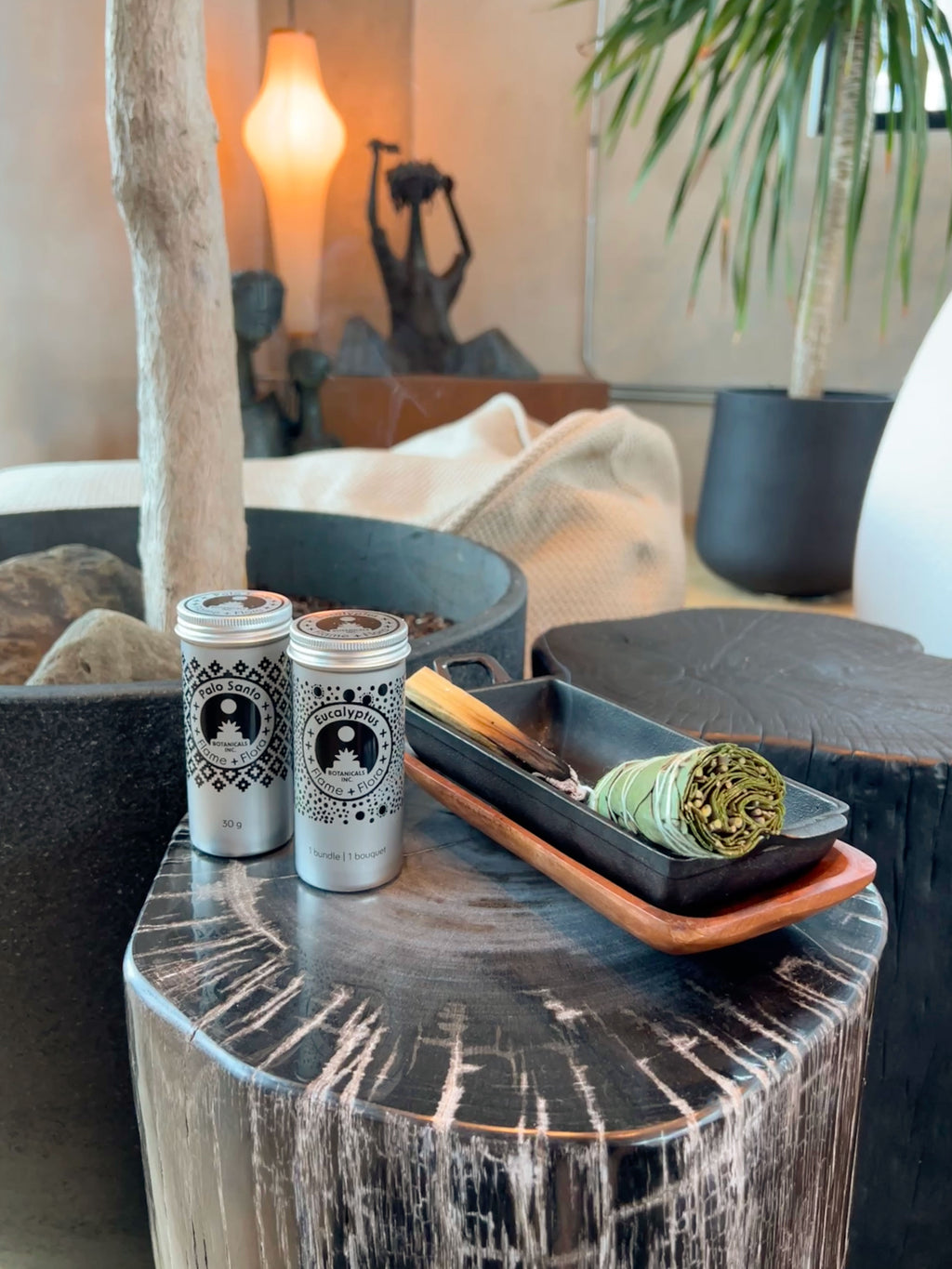 Two Flame and Flora cans with sit on a rustic dark wood side table next to a tray holding a bundle of sage smudge and a smouldering stick of Palo Santo, in a styled indoor setting with plants and warm ambient lighting.