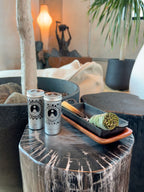 Two Flame and Flora cans with sit on a rustic dark wood side table next to a tray holding a bundle of sage smudge and a smouldering stick of Palo Santo, in a styled indoor setting with plants and warm ambient lighting.