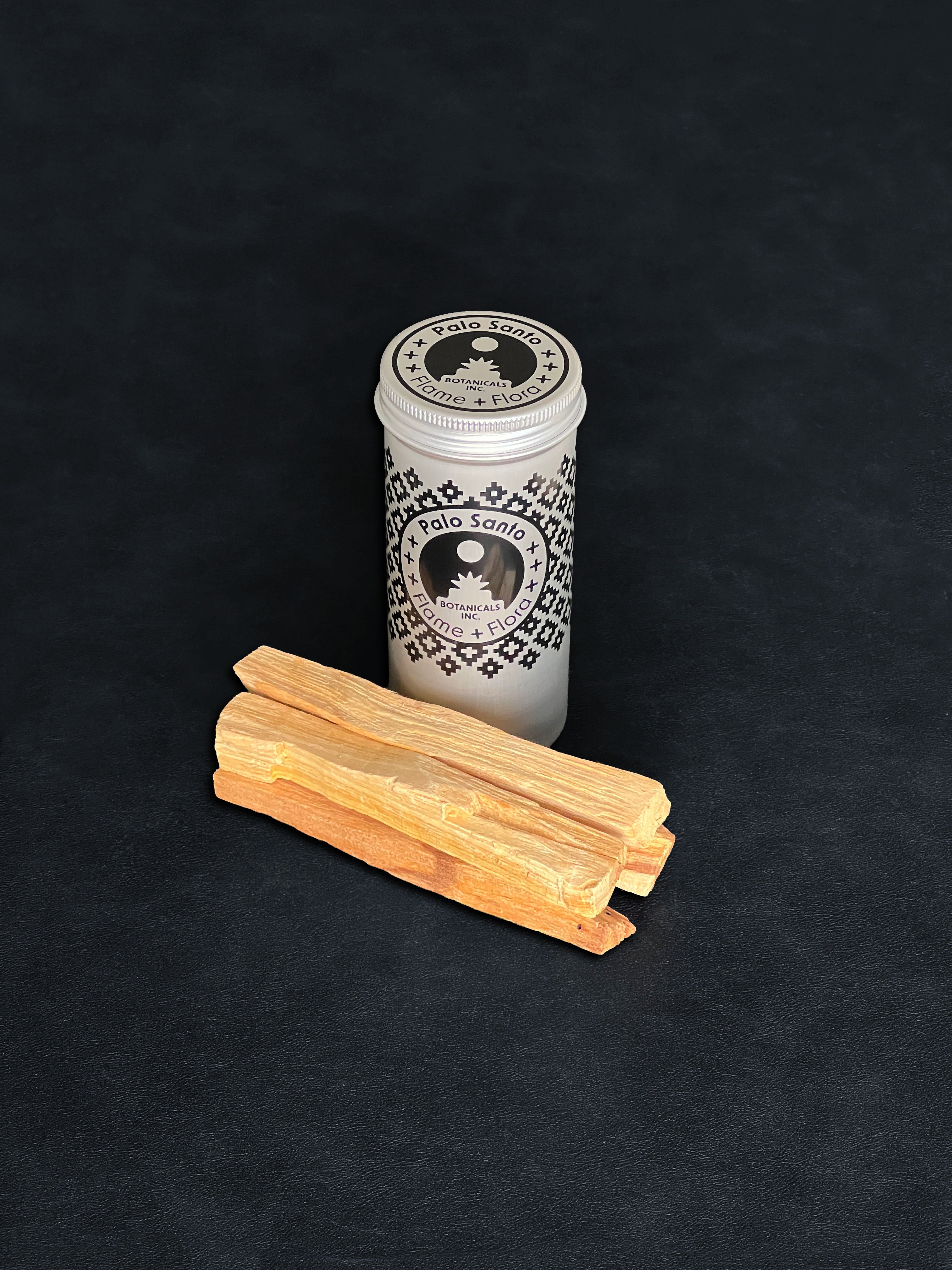 Palo Santo storage tin with black patterned design and four palo santo sticks displayed in the foreground on a dark background