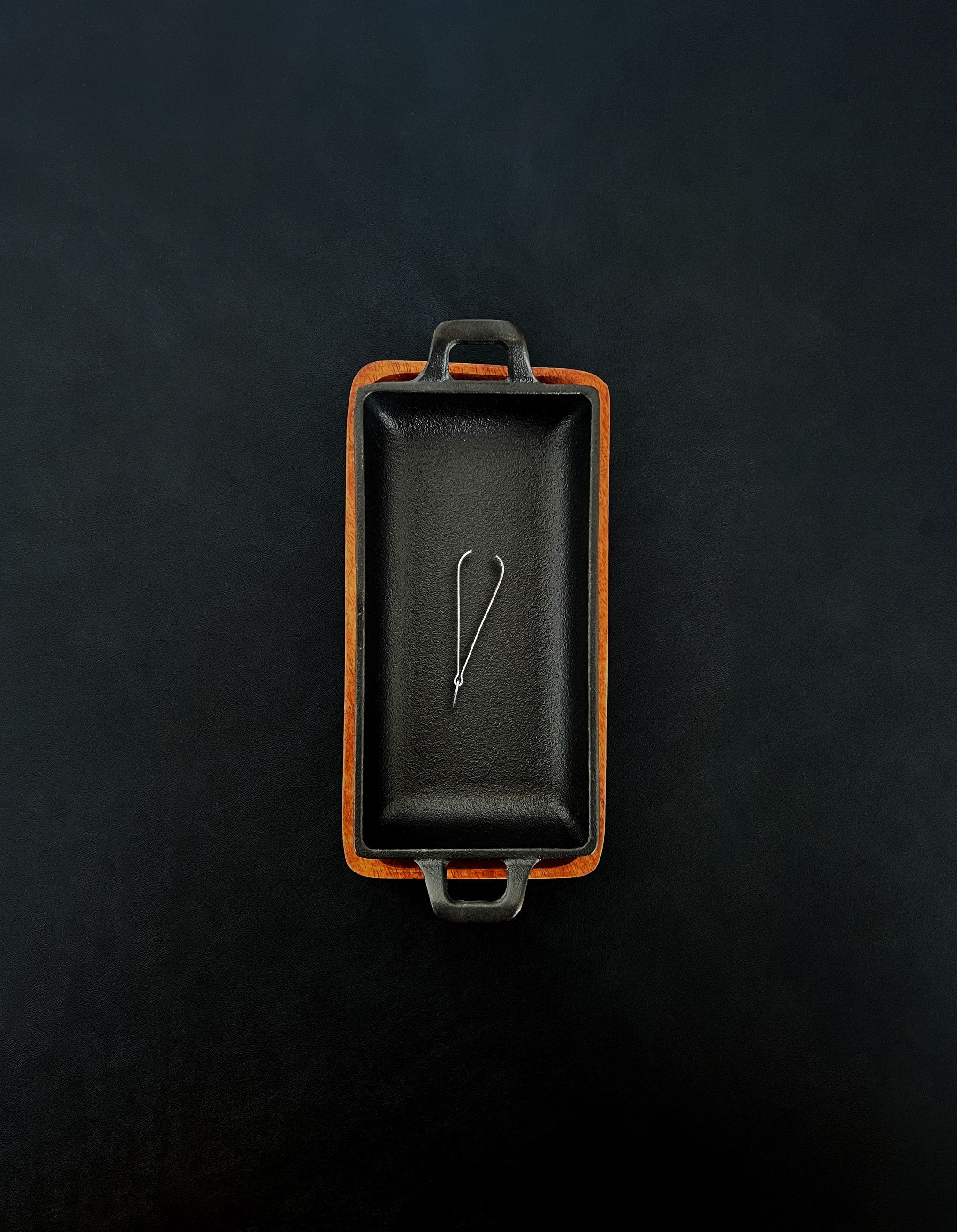 Bird’s-eye view of a rectangular black cast iron incense burner in a vertical orientation with compact metal tongs, nested in an acacia wood tray against a dark background