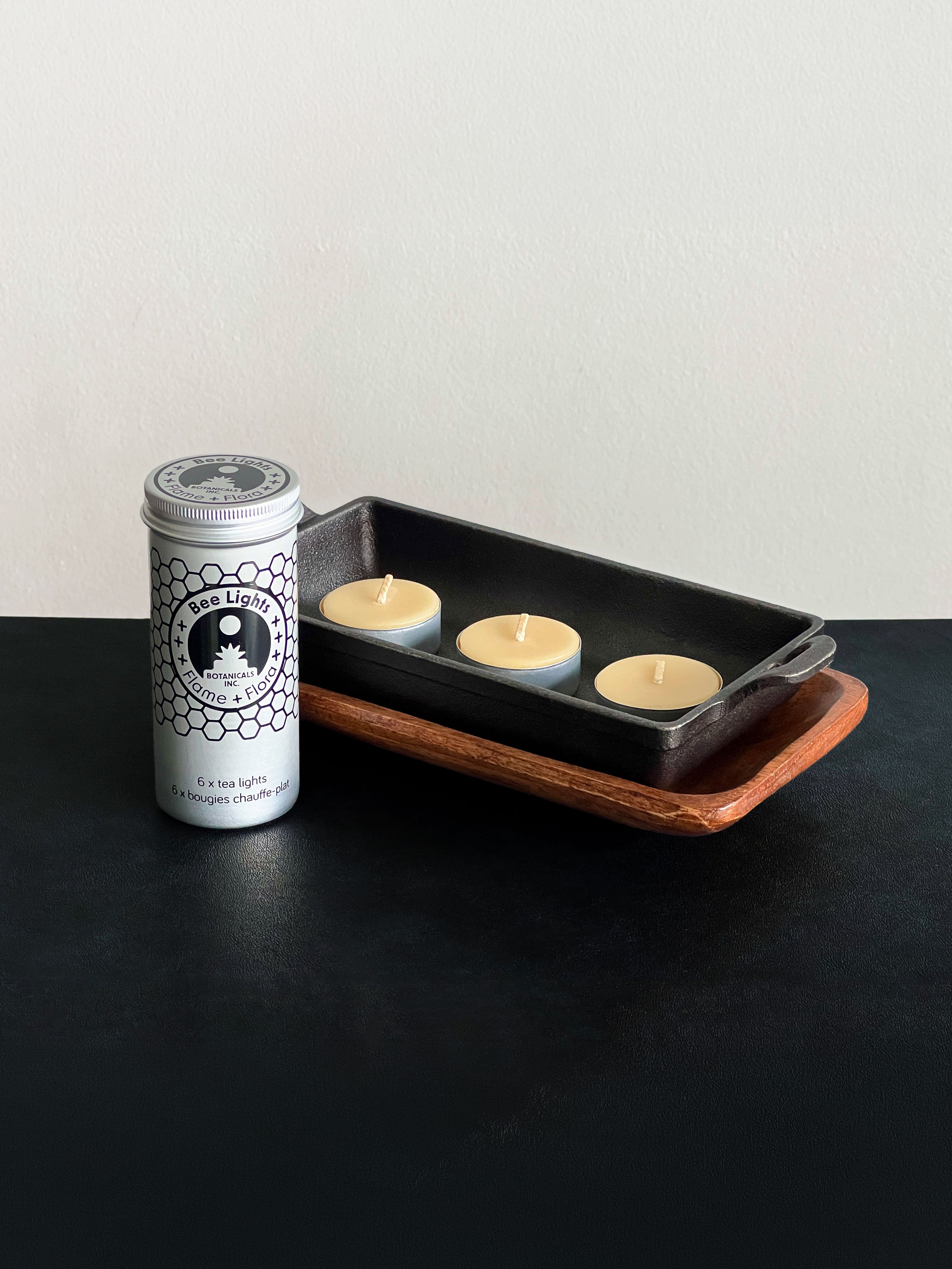 Beeswax tealight storage tin with black patterned design and three beeswax tealights in metal cups displayed with a cast iron incense burner and acacia wood tray set against a white background