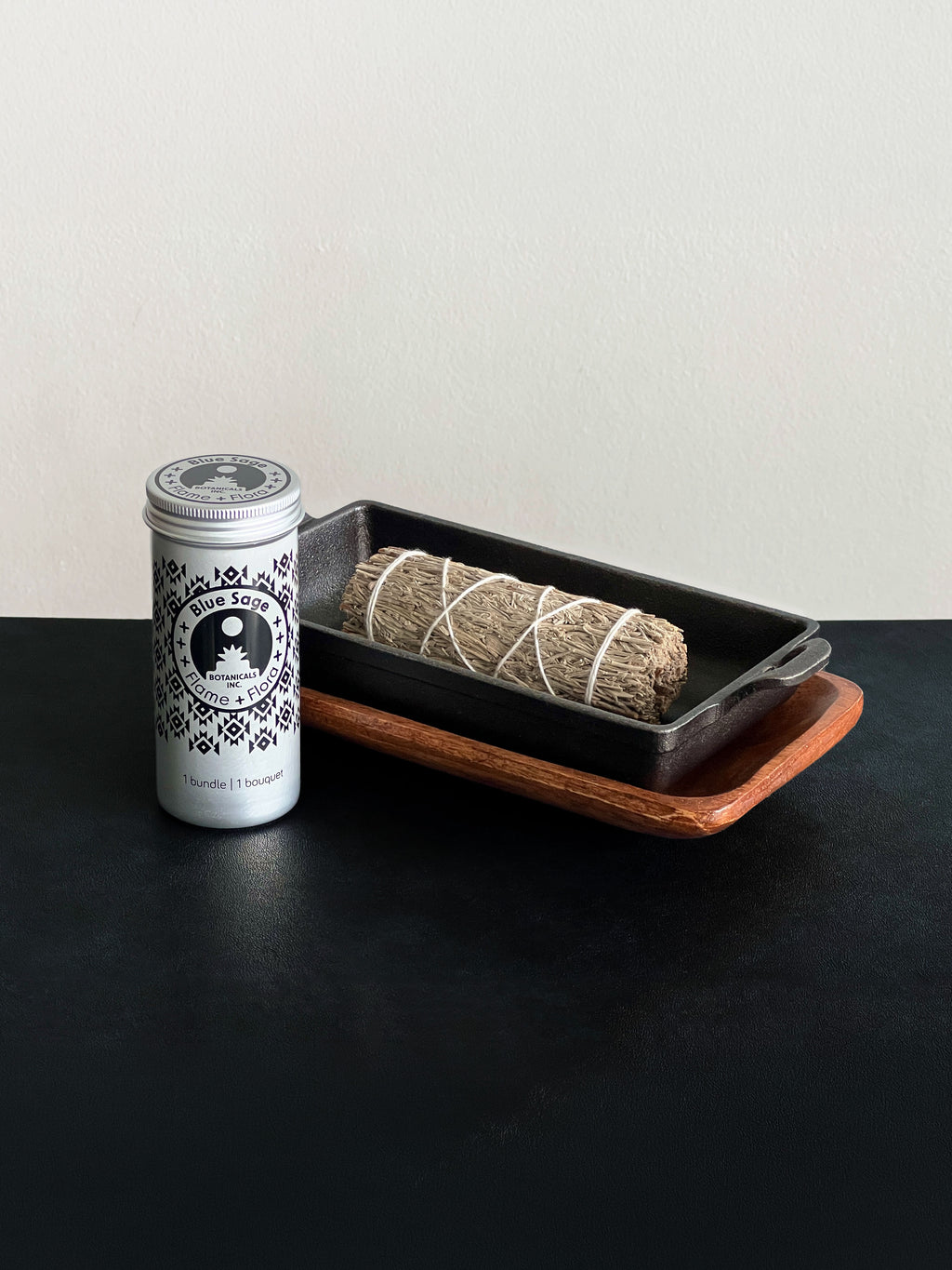Blue Sage storage tin with black patterned design and a bundle of Blue Sage tied with white cotton string displayed with a cast iron incense burner and acacia wood tray set on a black surface against a white background