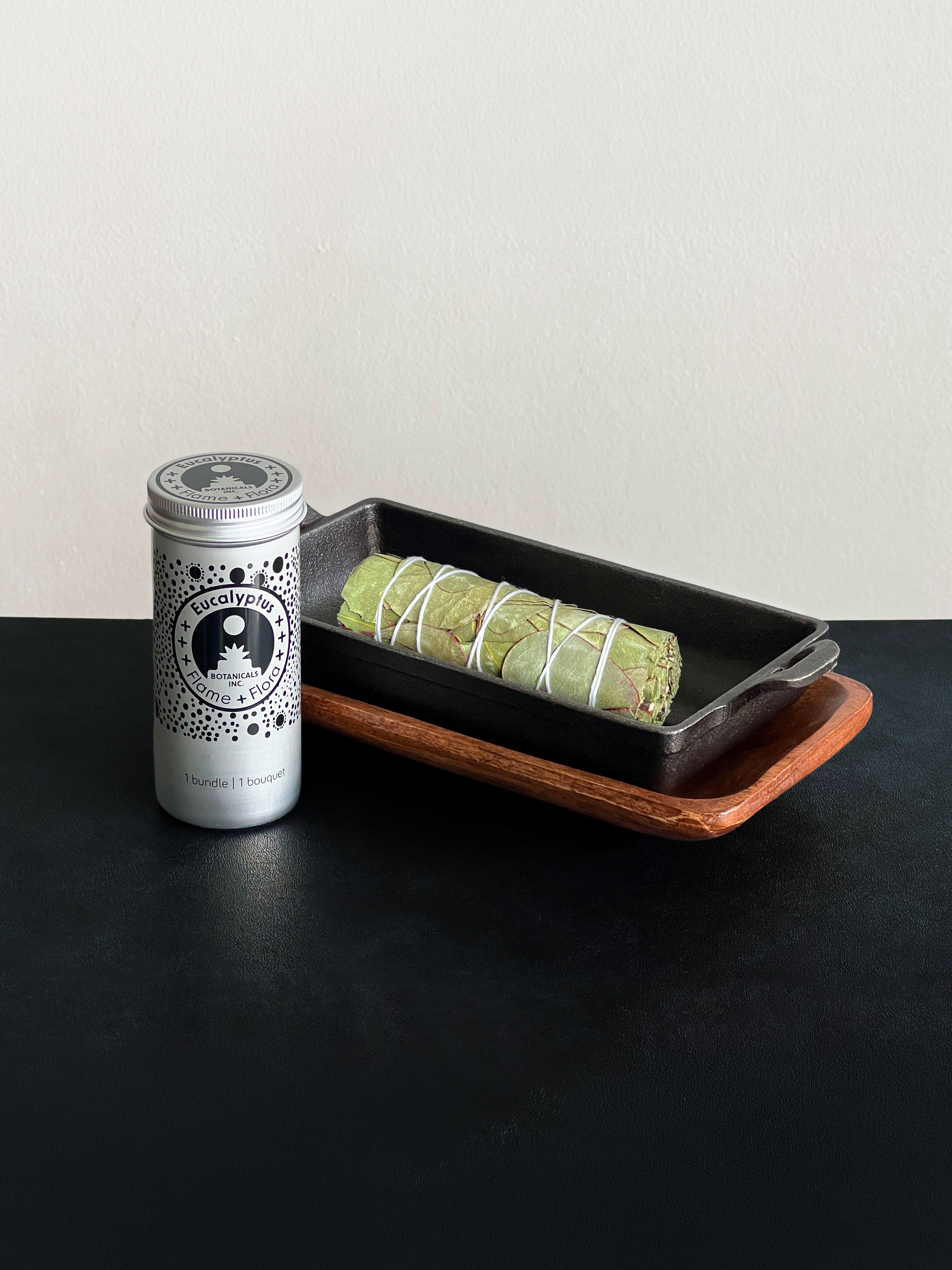 Eucalyptus storage tin with black patterned design and a bundle of Eucalyptus tied with white cotton string displayed with a cast iron incense burner and acacia wood tray set on a black surface against a white background