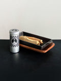 Palo Santo storage tin with black patterned design and Palo Santo sticks displayed with a cast iron incense burner and acacia wood tray set on a black surface against a white background
