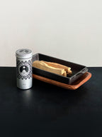 Palo Santo storage tin with black patterned design and Palo Santo sticks displayed with a cast iron incense burner and acacia wood tray set on a black surface against a white background
