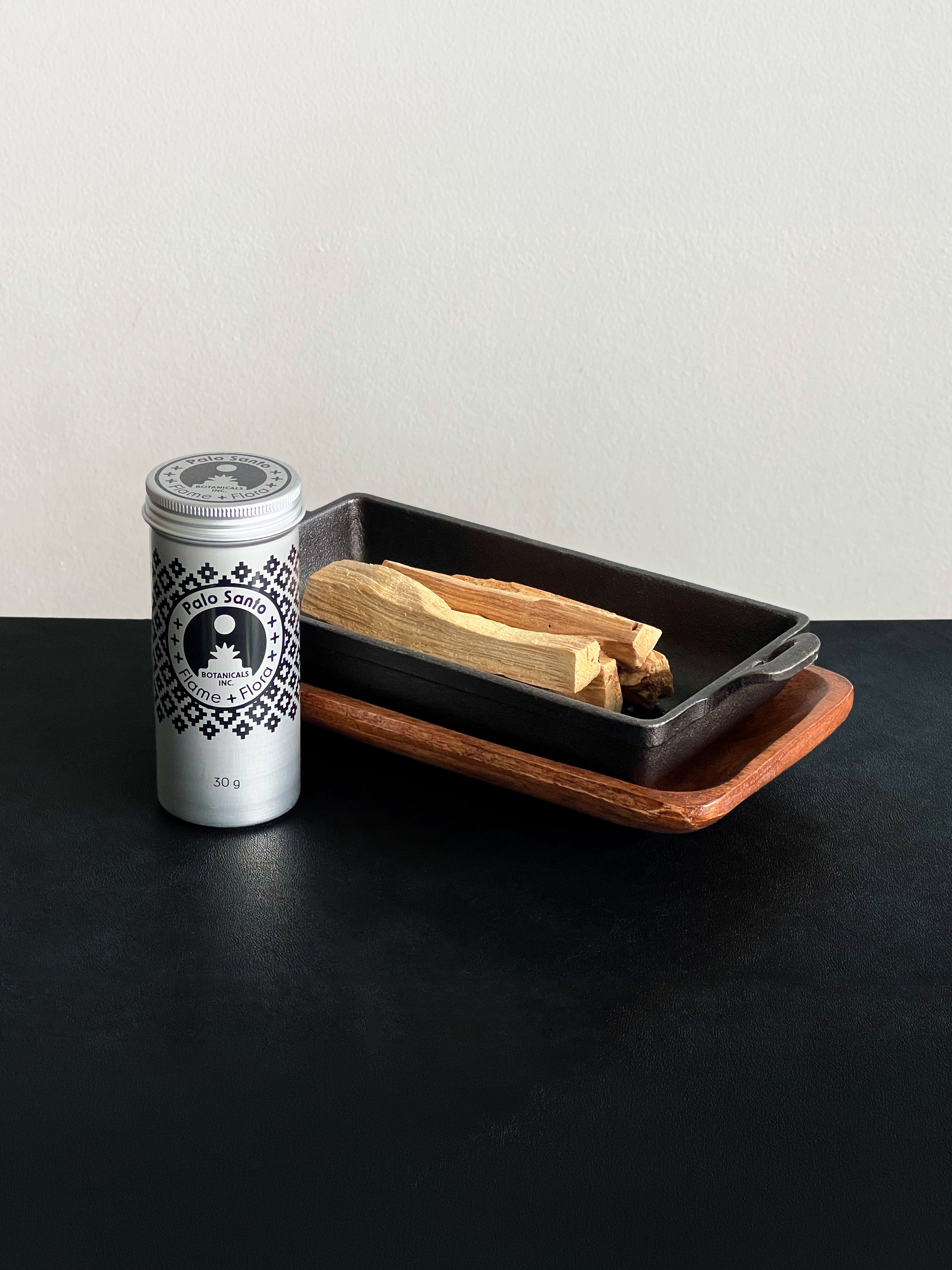 Palo Santo storage tin with black patterned design and Palo Santo sticks displayed with a cast iron incense burner and acacia wood tray set on a black surface against a white background