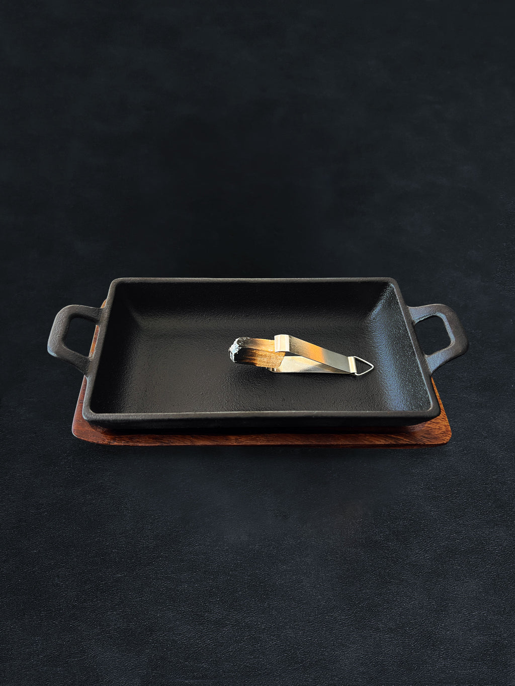 Rectangular black cast iron incense burner with acacia wood tray base holding metal tongs on a dark background