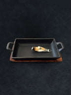 Rectangular black cast iron incense burner with acacia wood tray base holding metal tongs on a dark background