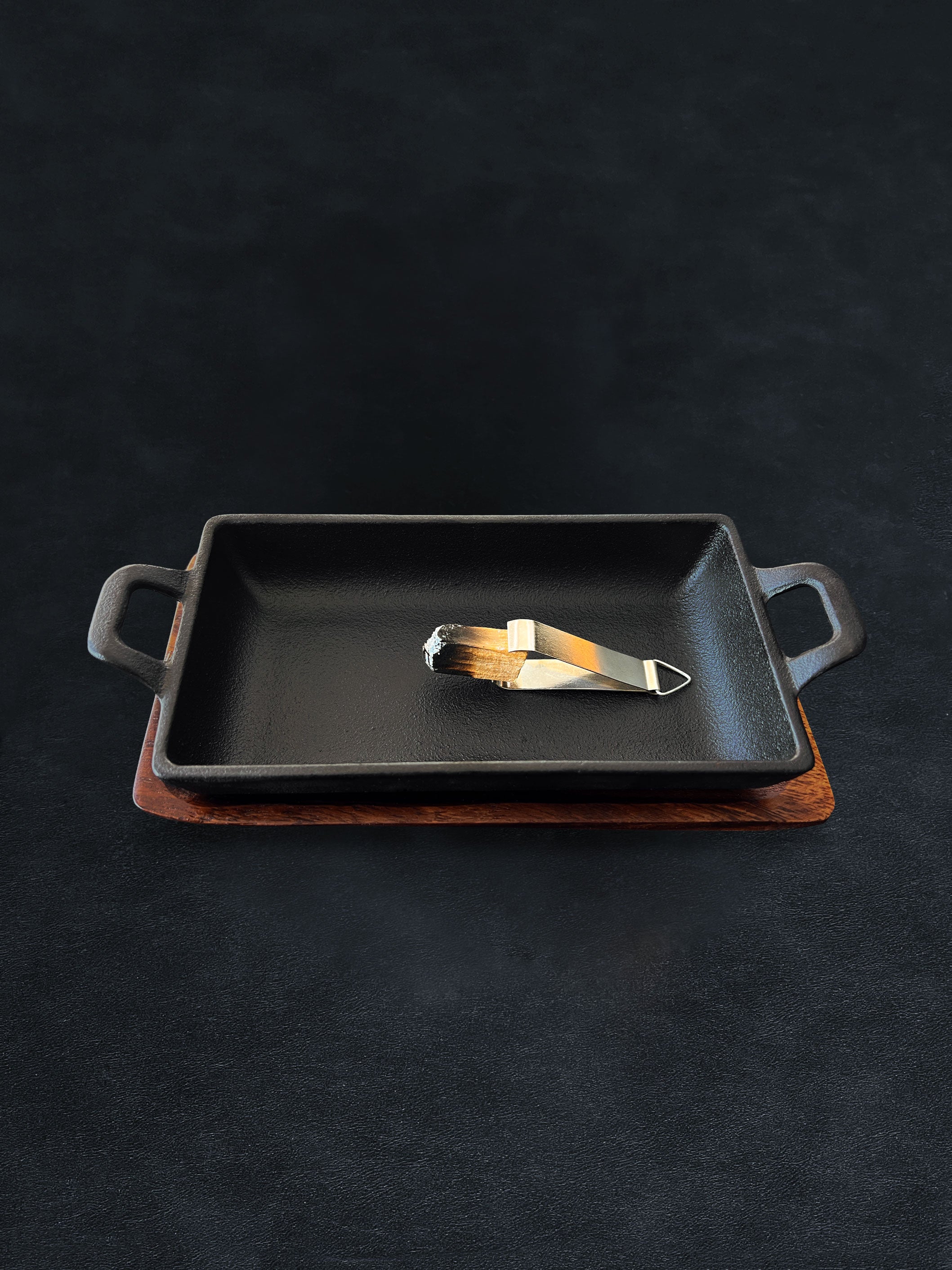 Rectangular black cast iron incense burner with acacia wood tray base holding metal tongs on a dark background