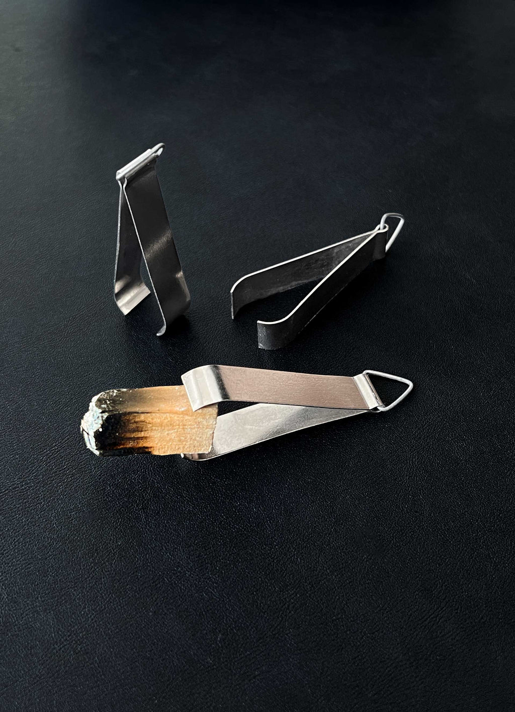 Three pairs of compact metal incense tongs against a dark background, one holding a charred piece of Palo Santo wood