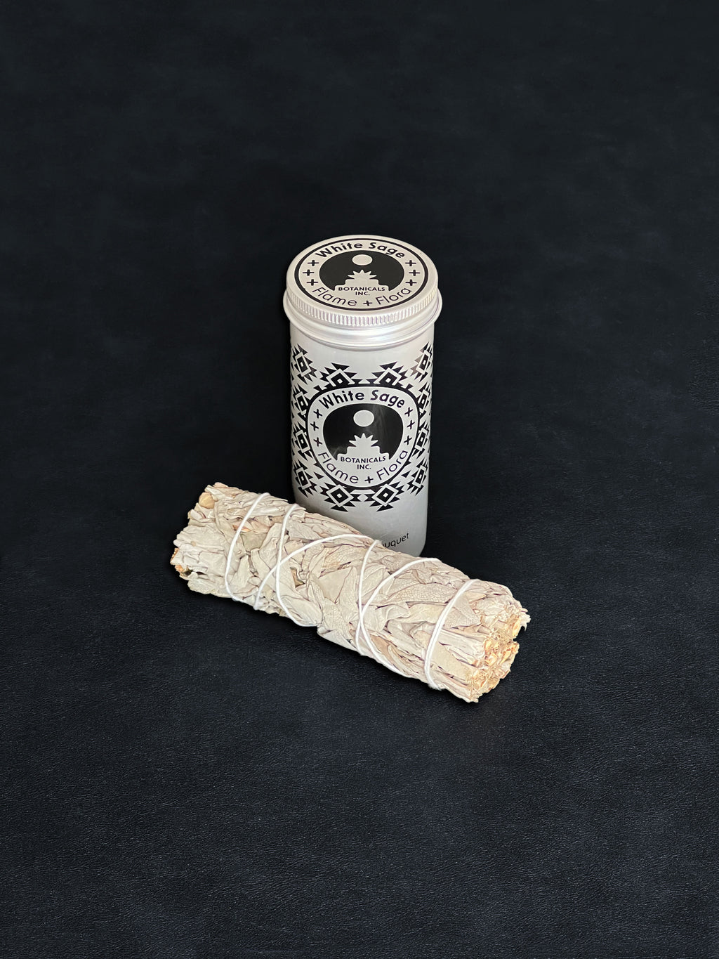 White Sage storage tin with black patterned design and a bundle of White Sage tied with white cotton string displayed in the foreground on a dark background