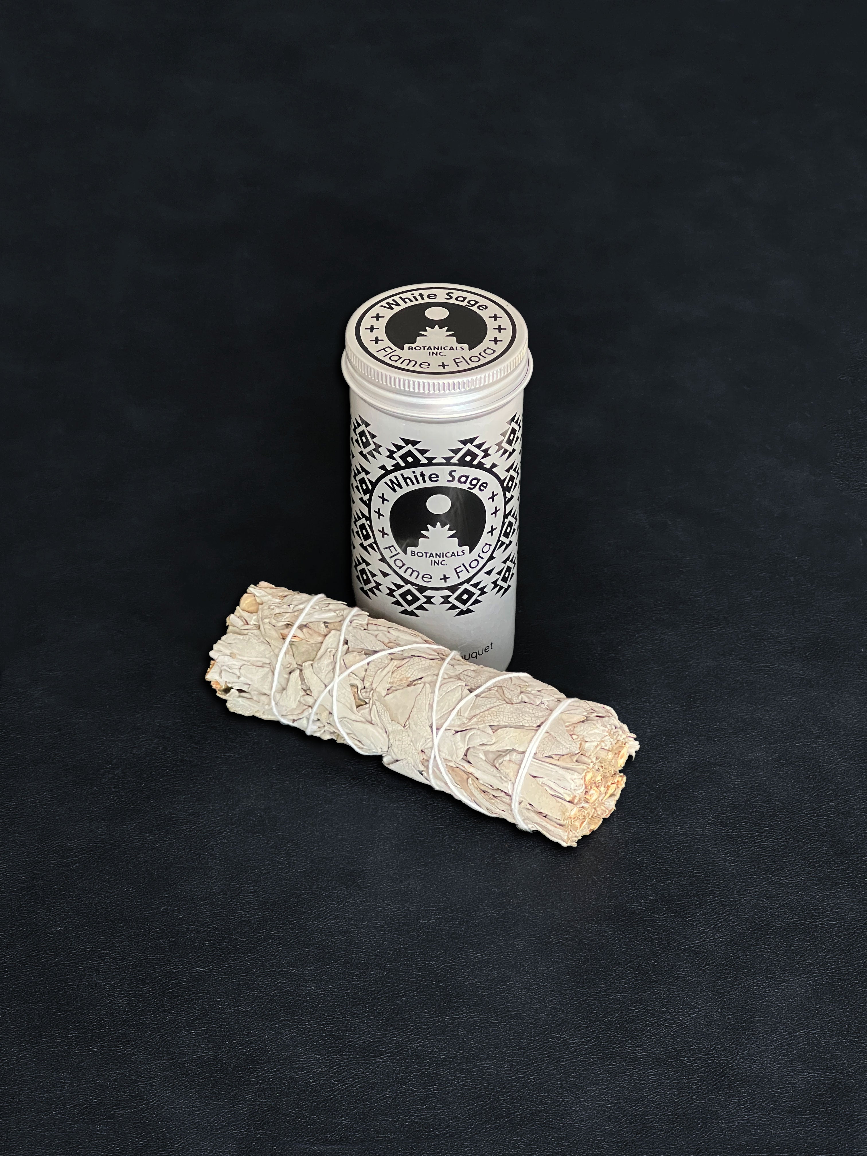 White Sage storage tin with black patterned design and a bundle of White Sage tied with white cotton string displayed in the foreground on a dark background