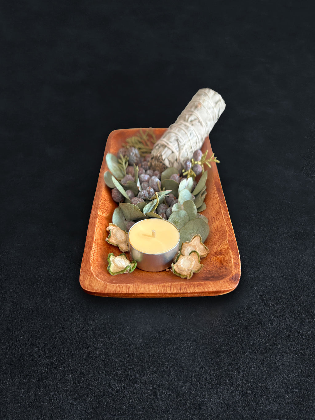 Bird’s-eye view of a rectangular acacia wood tray filled with loose botanicals and a white sage smudge stick positioned vertically on a dark surface against a white background