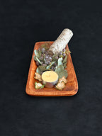 Bird’s-eye view of a rectangular acacia wood tray filled with loose botanicals and a white sage smudge stick positioned vertically on a dark surface against a white background