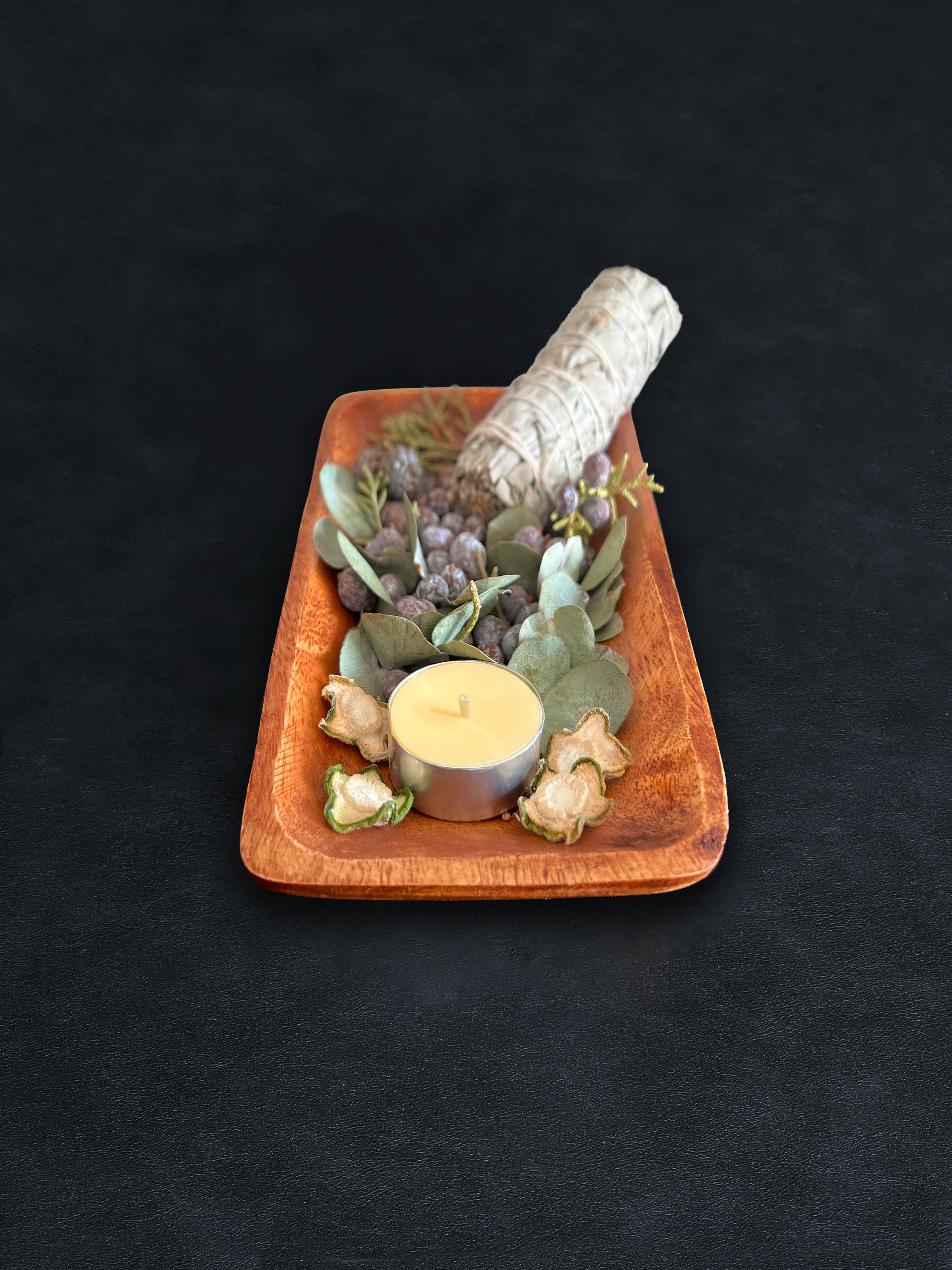 Bird’s-eye view of a rectangular acacia wood tray filled with loose botanicals and a white sage smudge stick positioned vertically on a dark surface against a white background