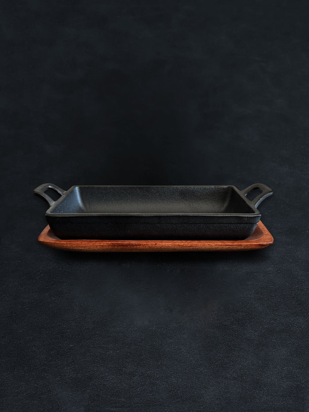 Side view of a rectangular black cast iron incense burner in a horizontal orientation with handles on both ends, nested in an acacia wood tray against a dark background
