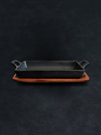 Side view of a rectangular black cast iron incense burner in a horizontal orientation with handles on both ends, nested in an acacia wood tray against a dark background