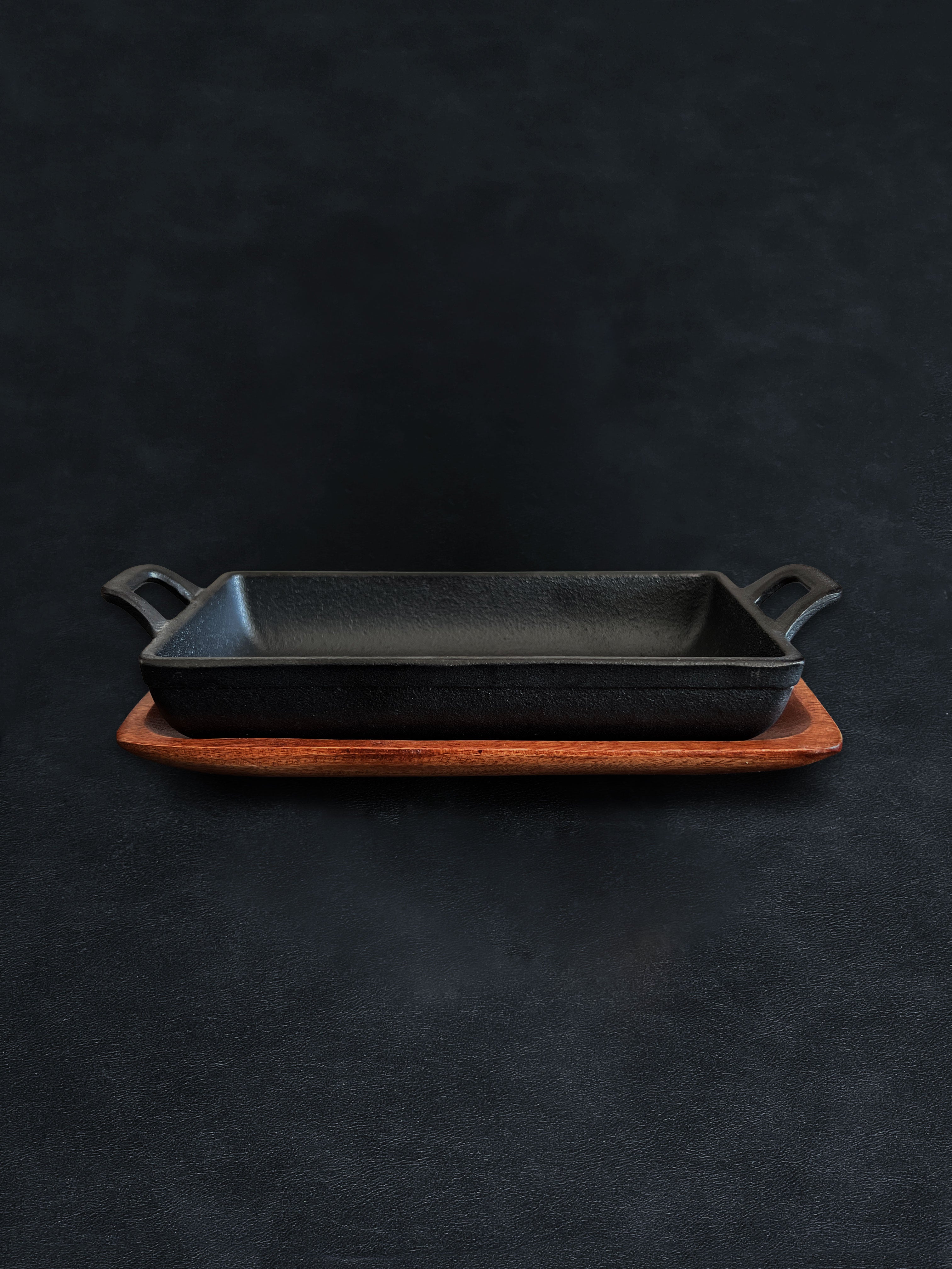 Side view of a rectangular black cast iron incense burner in a horizontal orientation with handles on both ends, nested in an acacia wood tray against a dark background