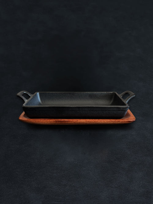 Side view of a rectangular black cast iron incense burner in a horizontal orientation with handles on both ends, nested in an acacia wood tray against a dark background