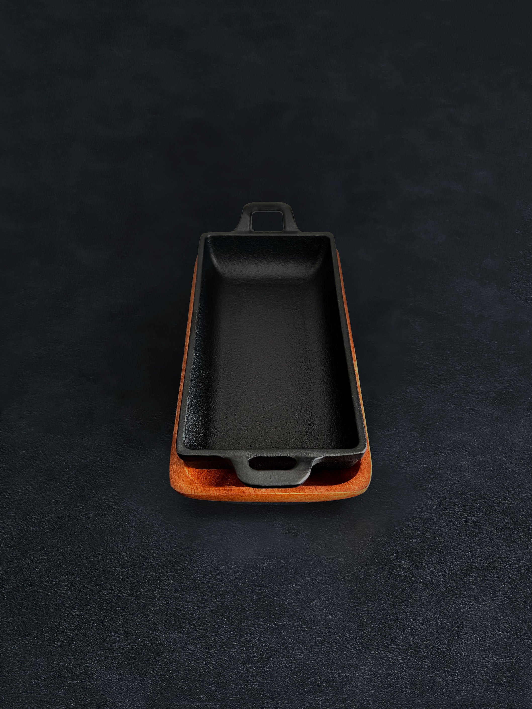 Bird’s-eye view of a rectangular black cast iron incense burner in a vertical orientation with handles on both ends, nested in an acacia wood tray on a dark surface against a white background